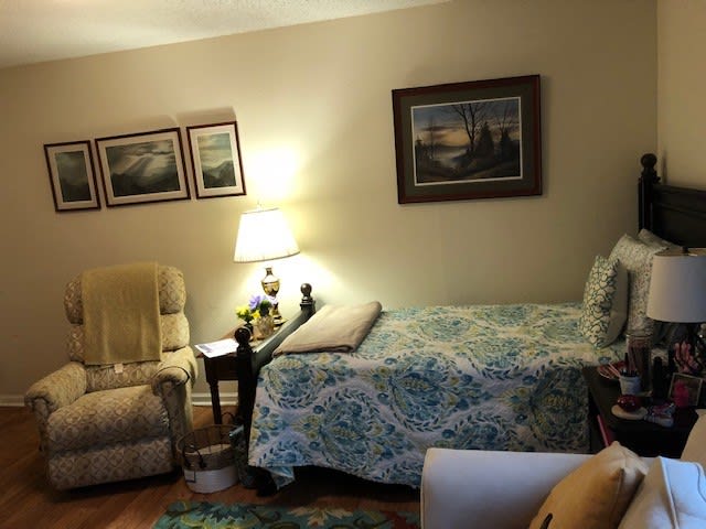 A cozy living room with a bed, reclining chair, lamp, and wall art.