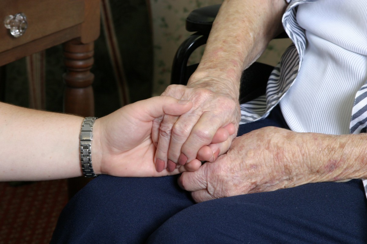 An older adult's hand is gently held by a younger person, symbolizing support and compassion.