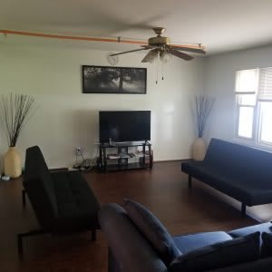 A spacious living room with modern decor, featuring a television, multiple sofas, a ceiling fan, and large windows.