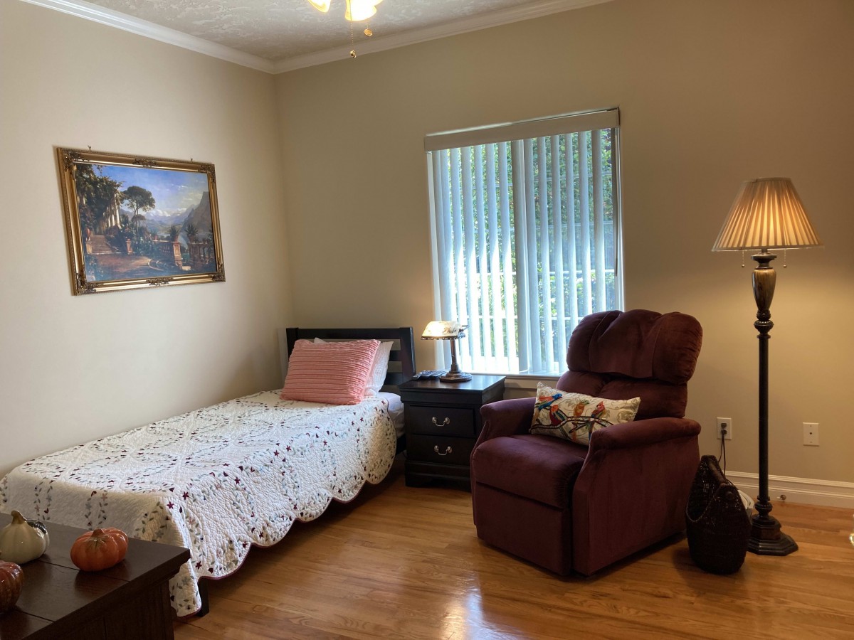 A cozy bedroom with a single bed, an armchair, and a lamp beside a window.