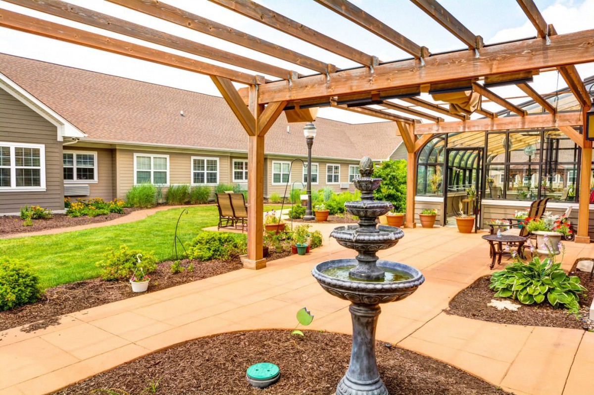 A tranquil garden area featuring a tiered fountain under a wooden pergola, surrounded by well-maintained landscaping and outdoor seating.