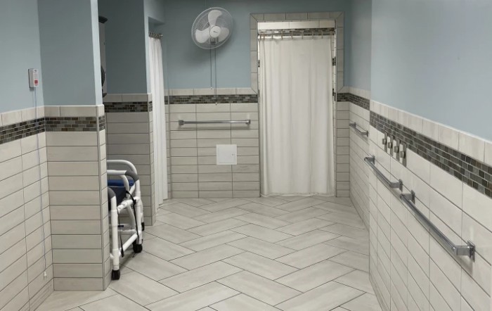 A hospital corridor with a wheelchair, tiled walls and floor, handrails, and a door at the end.