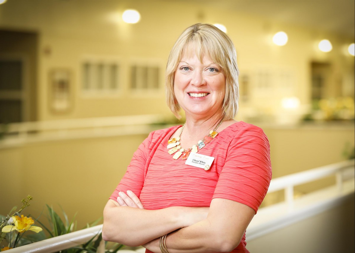 A smiling woman with blonde hair wearing a red dress and a name tag stands indoors.