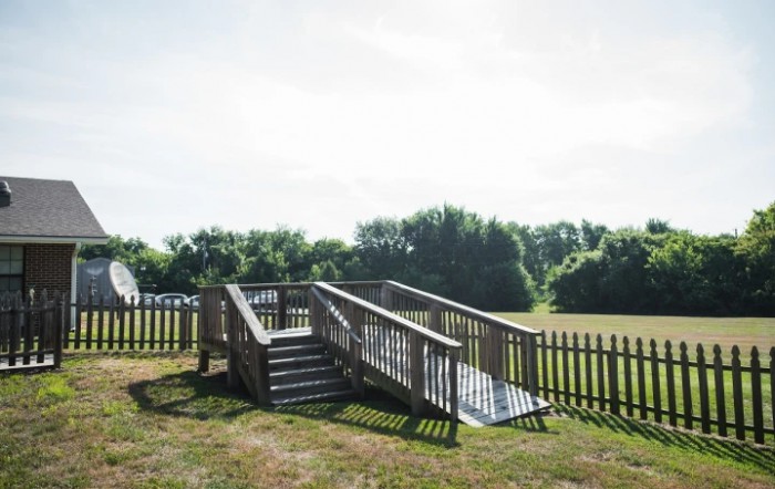 A wooden accessibility ramp and stairs leading to a fenced grassy area on a sunny day.
