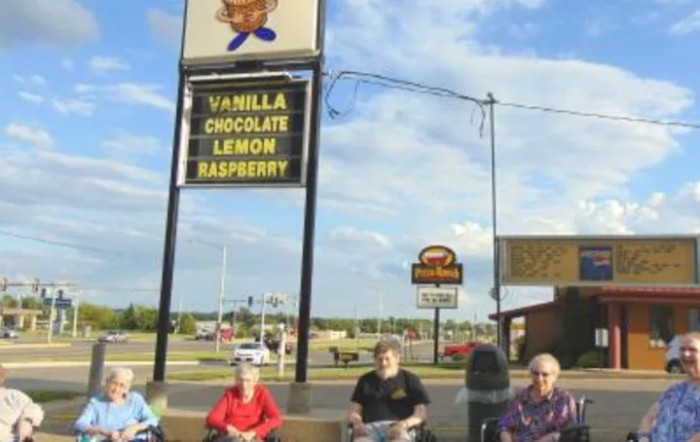 A group of people sit outdoors near an ice cream shop sign listing flavors: vanilla, chocolate, lemon, and raspberry.