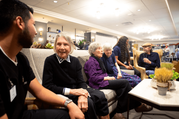 A group of people, including seniors, are sitting and chatting indoors on a couch.