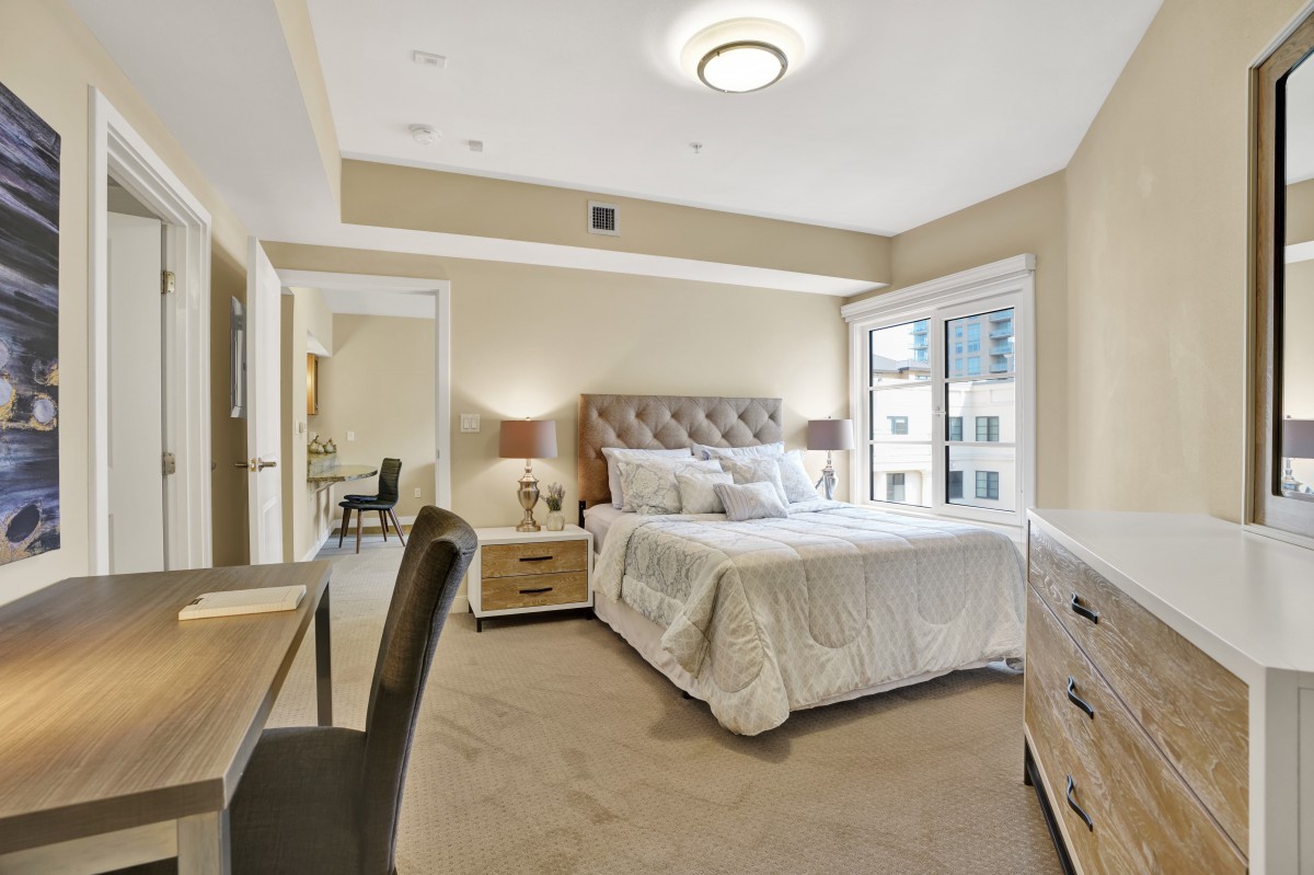 A modern bedroom featuring a plush bed with neutral tones, ample natural light from a large window, and elegant furniture.