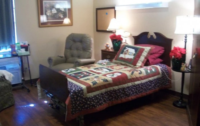 A cozy bedroom featuring a bed with a colorful quilt, an armchair, and a lamp on the bedside table.