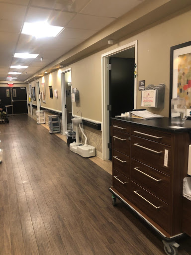 A view down a well-lit hallway in a medical facility, showcasing doors and furniture typical of a hospital setting.
