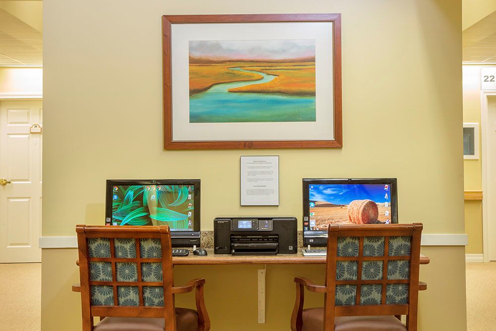 A computer workstation setup with two monitors, a printer, and chairs, featuring artwork on the wall.