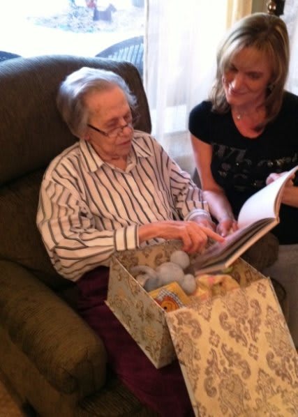 An elderly person and a younger woman sit together, looking at a book in a cozy setting.