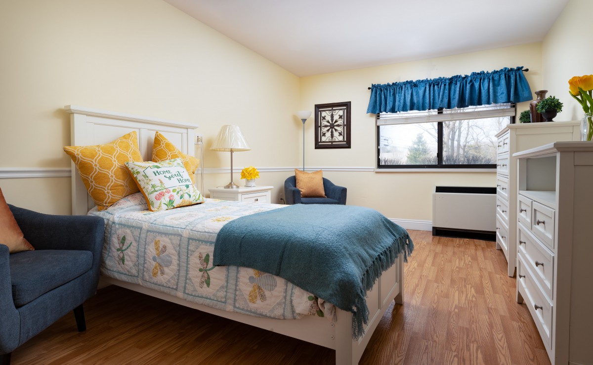 Cozy bedroom with a single bed featuring colorful pillows and a decorative quilt, complemented by a window with blue curtains and a wooden floor.