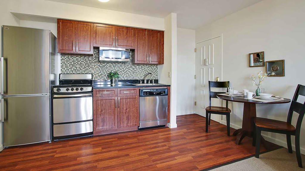 A modern kitchen with stainless steel appliances, wood cabinetry, and a small dining table in an open layout.
