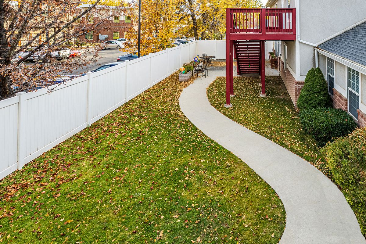 A backyard with a winding concrete walkway, bordered by a white fence and featuring autumn foliage.