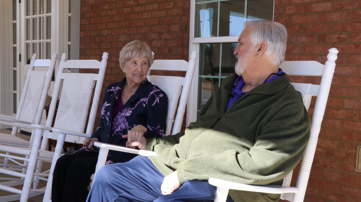 An elderly couple sitting on rocking chairs outdoors, engaged in conversation.