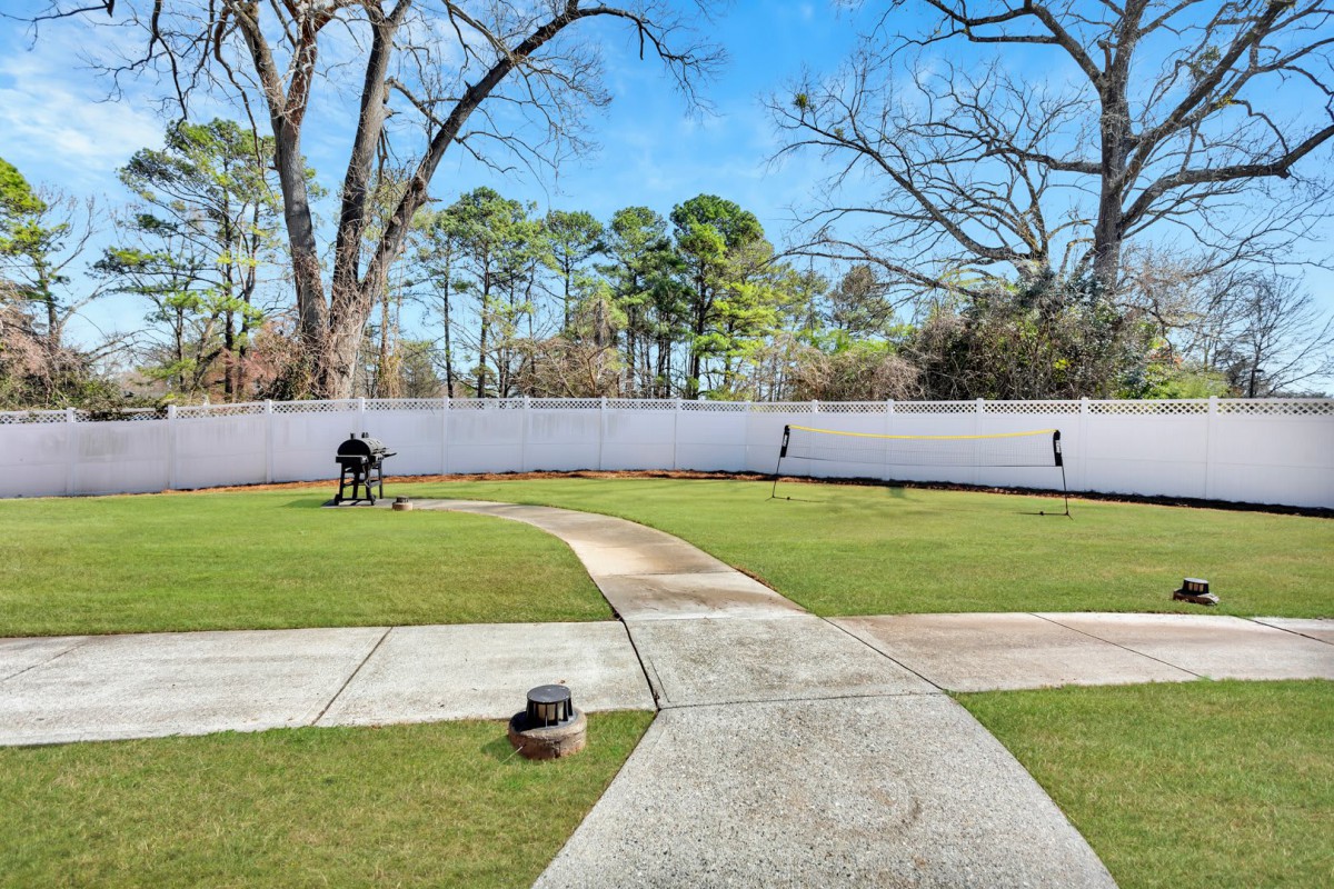 A tidy backyard with a concrete pathway, green lawn, and tall trees behind a white fence.