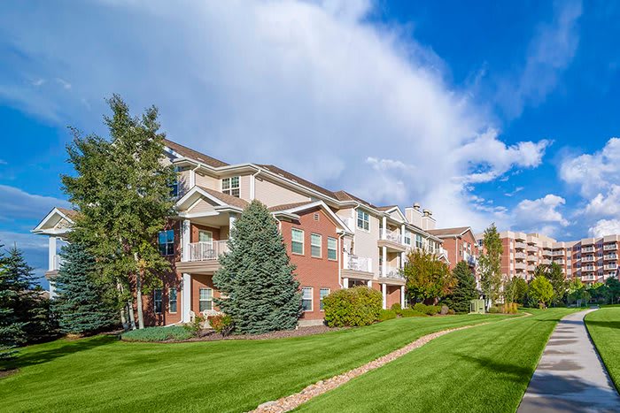 A scenic view of a residential apartment building surrounded by lush grass and trees under a partly cloudy sky.
