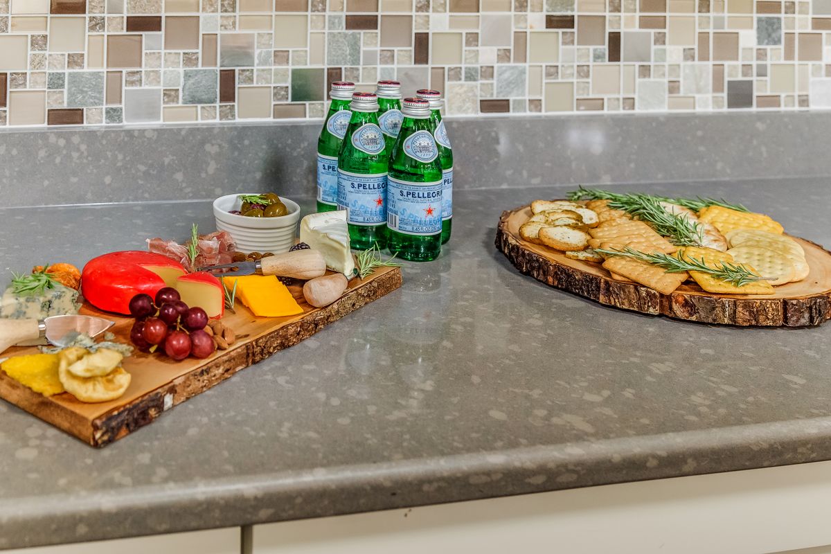 A kitchen counter features a charcuterie board with assorted cheeses, crackers, and grapes, alongside bottles of sparkling water.