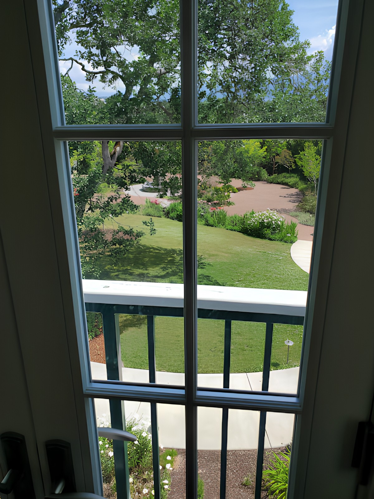 A view of a garden and trees through a window with a balcony railing.