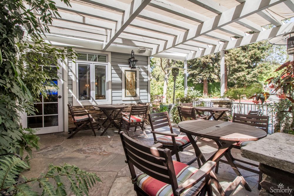 A serene outdoor patio area with wooden tables and chairs under a pergola, surrounded by lush garden greenery.