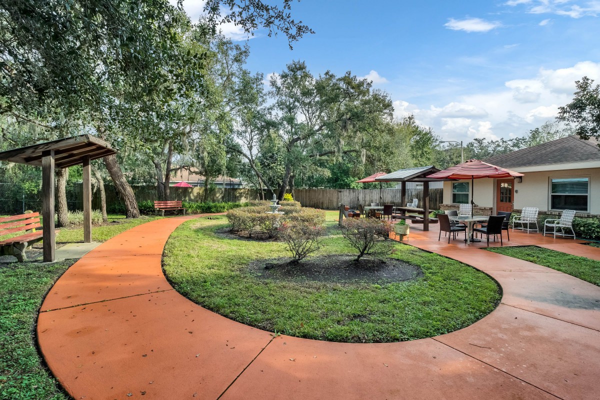 A landscaped garden with a circular pathway, surrounded by trees and shrubs, adjacent to a house with a patio.