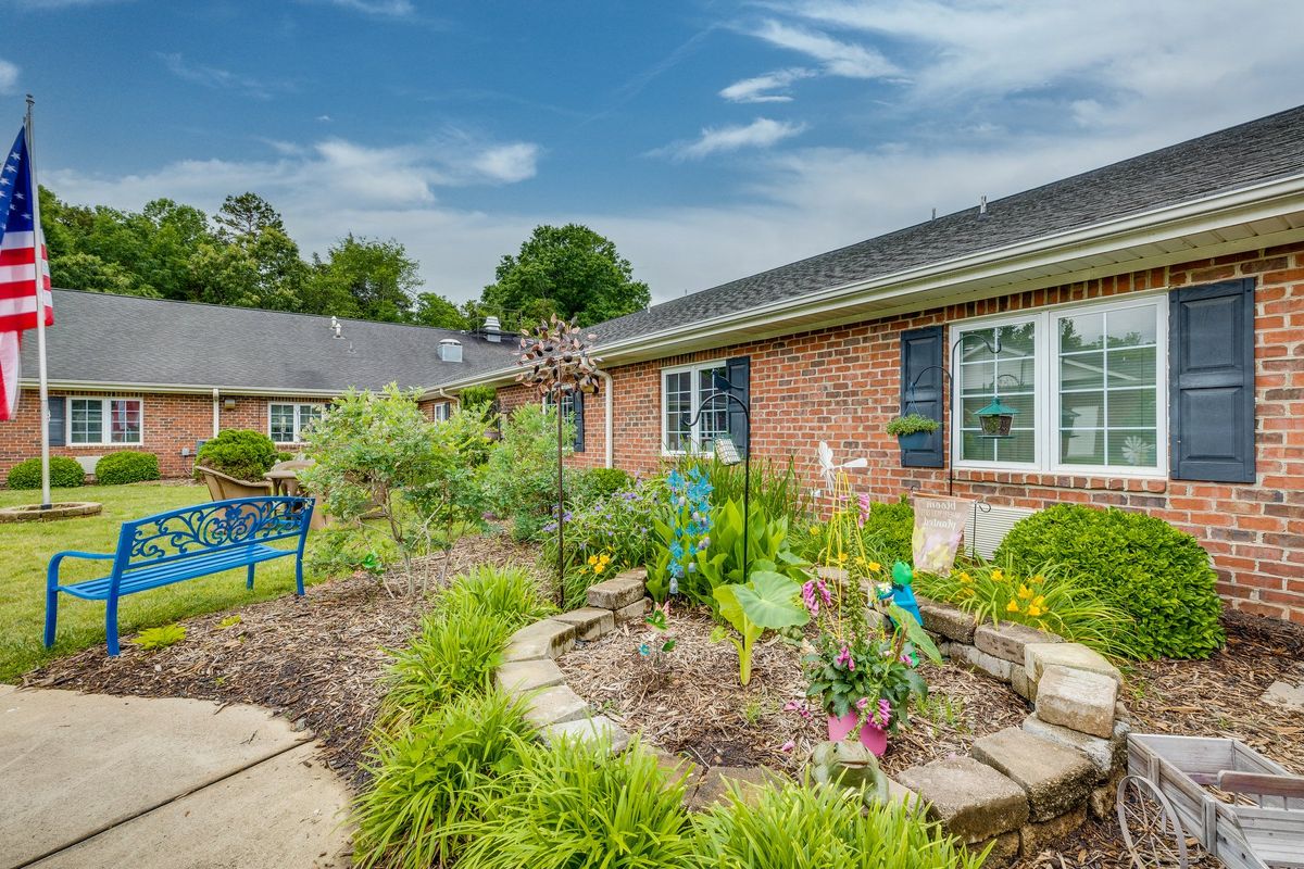 A well-maintained garden in front of a brick house, featuring a variety of plants and a decorative bench.