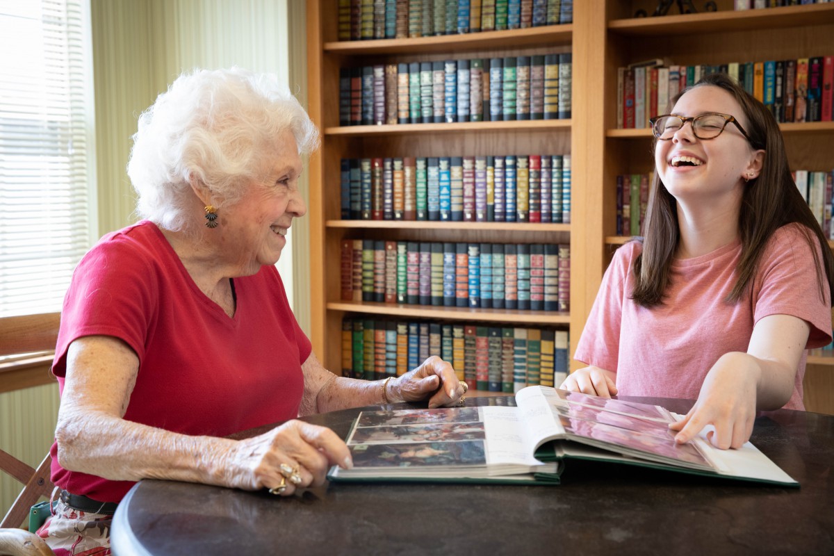 An elderly woman and a young woman share a moment looking through a photo album in a library.