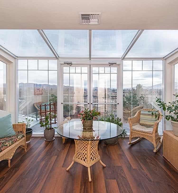 Bright sunroom with wicker furniture and abundant natural light, featuring glass walls and a wooden floor.