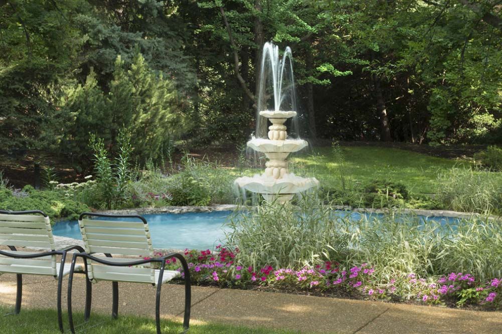 A peaceful garden scene with a flowing fountain and a bench surrounded by lush greenery and colorful flowers.