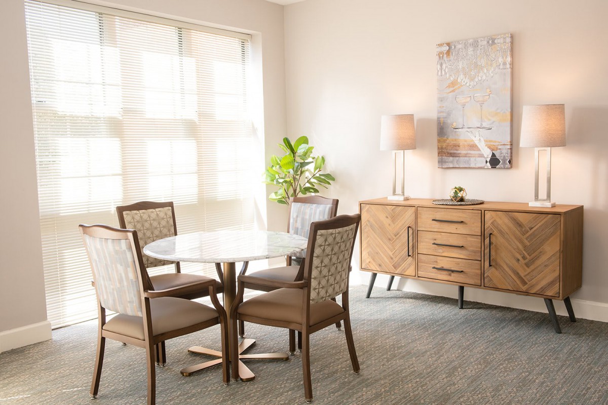 A bright dining room featuring a round table with four chairs, a sideboard with a lamp and plant, and wall art in a modern interior design.