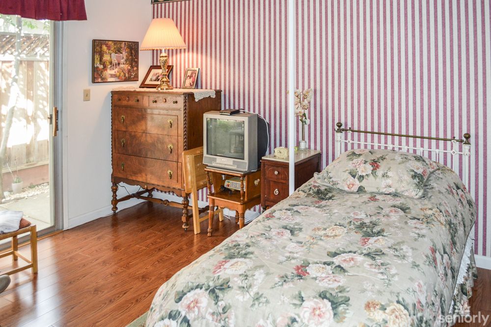 A cozy bedroom with floral bedding, featuring antique furniture and a vintage television against striped wallpaper.