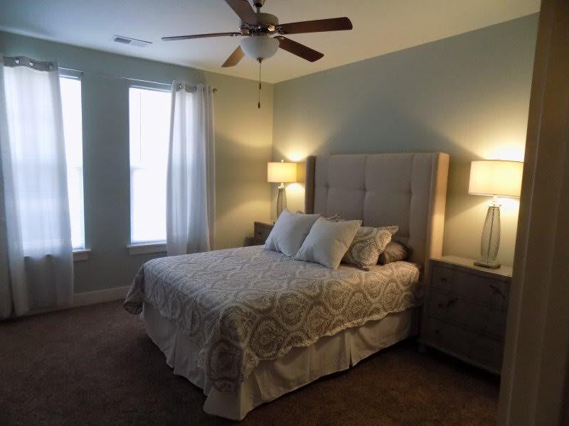 A cozy bedroom with a neatly made bed, two lamps on nightstands, and large windows with curtains.