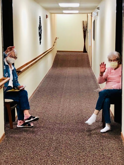 Two people wearing masks engage in a socially-distanced conversation in a hallway.