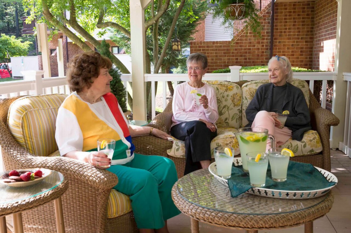 Three elderly women seated on a porch engage in conversation while enjoying refreshments.