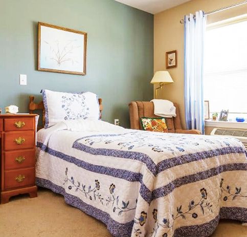 A cozy bedroom with a single bed and a quilt, featuring a nightstand, armchair, lamp, and window.