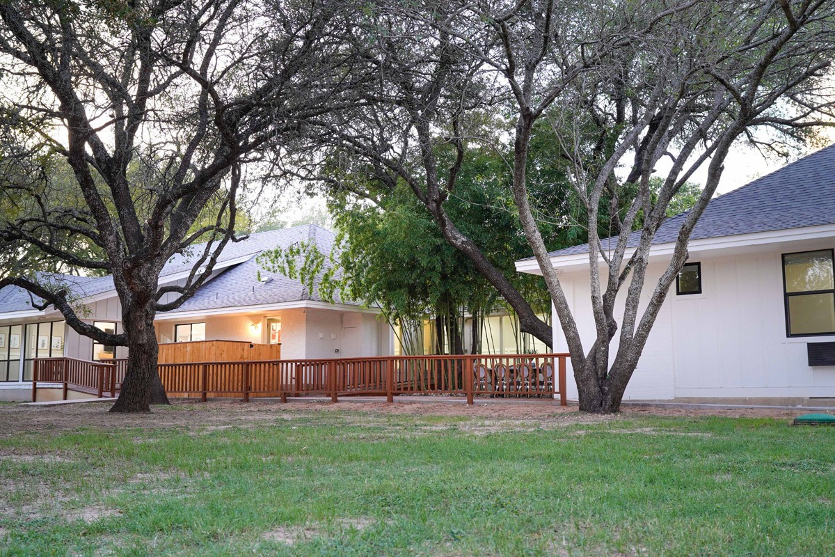 A tranquil backyard featuring a white house with a wooden deck surrounded by lush trees and grass.