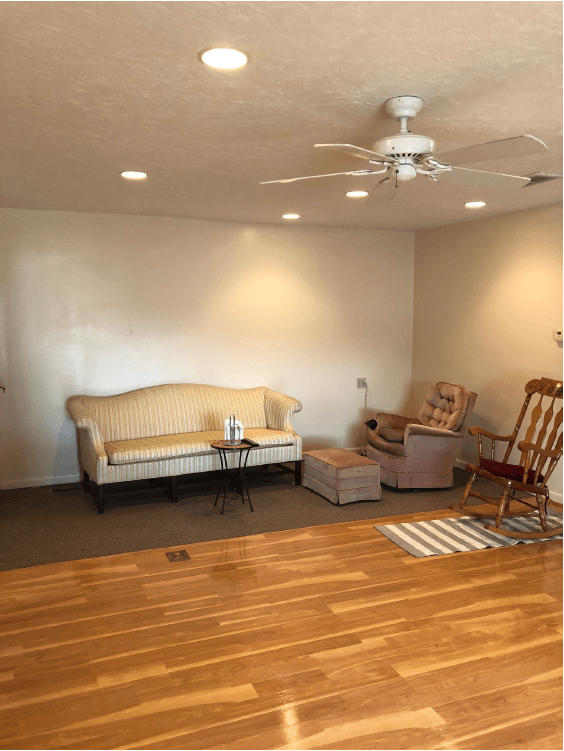 A cozy living room with a sofa, chairs, and a rocking chair on a wooden floor, featuring recessed lighting and a ceiling fan.