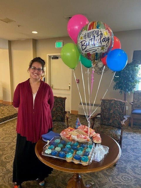 A woman stands next to a table with birthday balloons, cupcakes, and a cake.