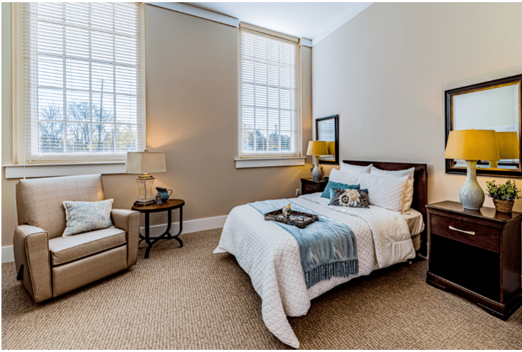 A cozy and well-lit bedroom featuring a comfortable bed with decorative pillows, a bedside table with a lamp, and an armchair in the corner.