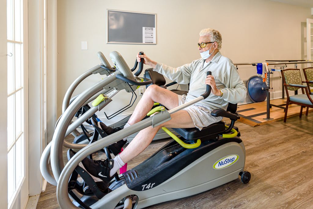 An elderly person exercising on a recumbent bike while wearing a mask in a gym setting.