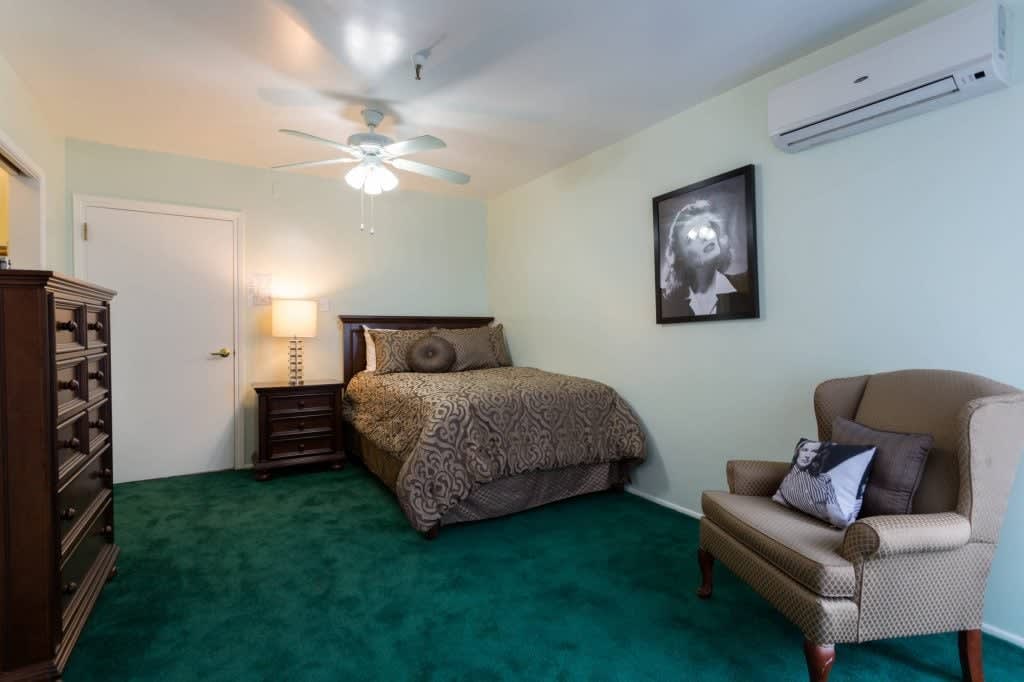 A cozy bedroom featuring a bed, armchair, and artwork on the wall with green carpet flooring.
