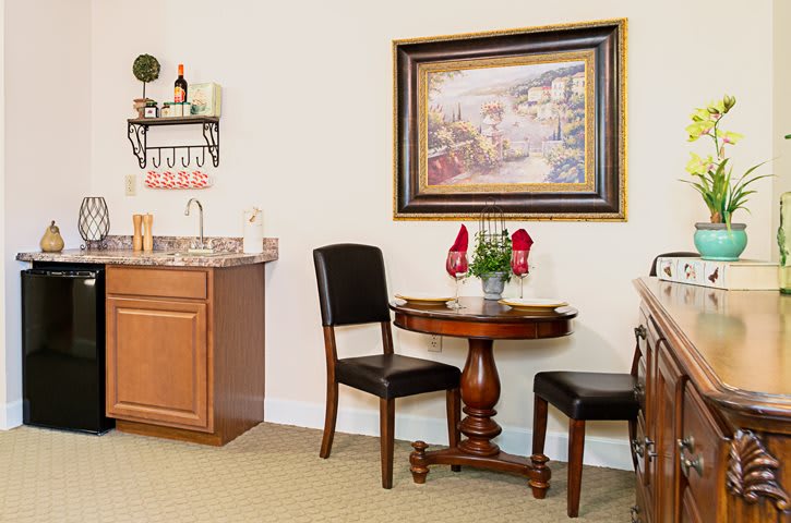 A cozy dining area with a round table set for two, complemented by a kitchenette and a decorative painting on the wall.