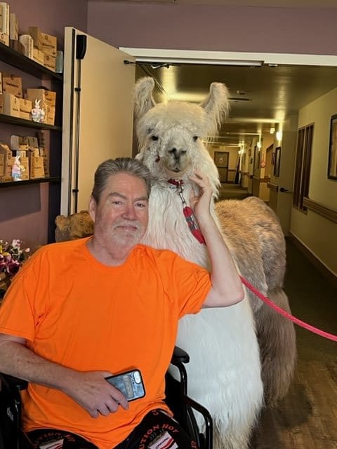 A man in an orange shirt smiles while petting a llama indoors.