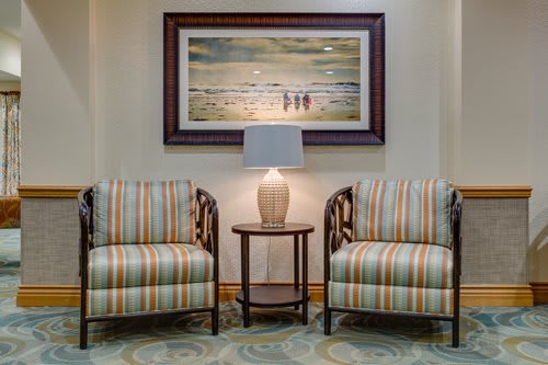 A cozy seating area with two striped chairs and a table topped with a lamp, framed by a seascape painting in the background.