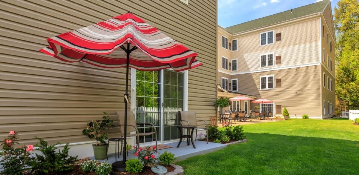 A cozy outdoor patio area with a red and white striped umbrella, surrounded by a lush green lawn next to a building.
