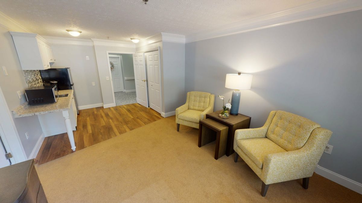 A cozy living room with two yellow chairs, a small table with a lamp, and a blend of carpet and hardwood flooring.