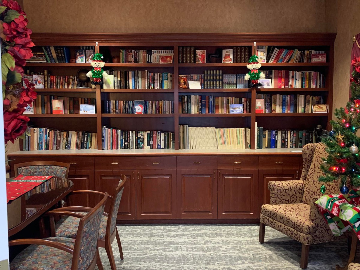 A cozy library room with a large wooden bookshelf filled with books, adorned by green lamps and accompanied by comfortable chairs.