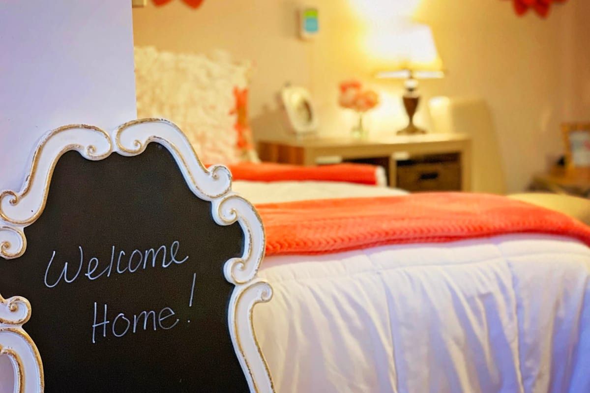 A cozy bedroom with a 'Welcome Home!' sign and warm lighting.
