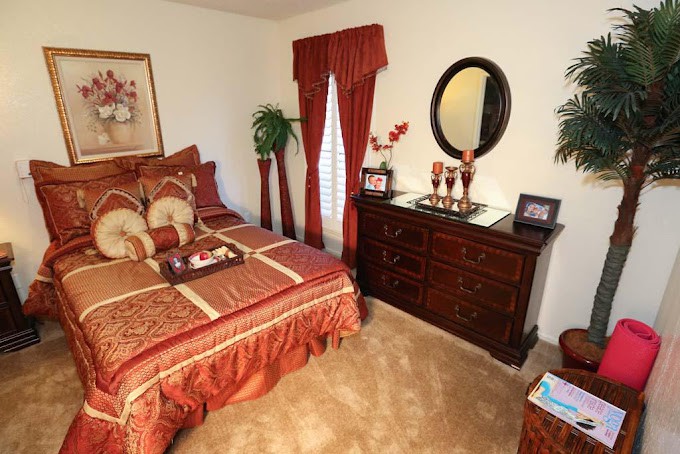 A neatly decorated bedroom with matching red and gold accents, featuring a bed, dresser, mirror, and potted plants.