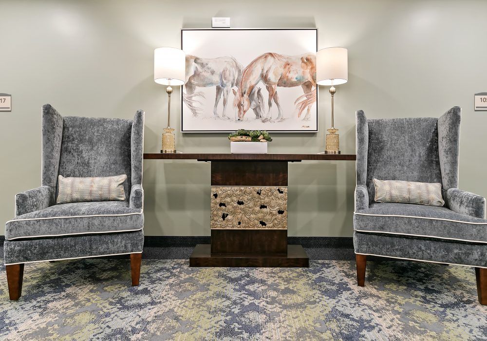 A cozy lobby area featuring two grey armchairs, a console table with drawers, decorative lamps, and a horse-themed painting on the wall.